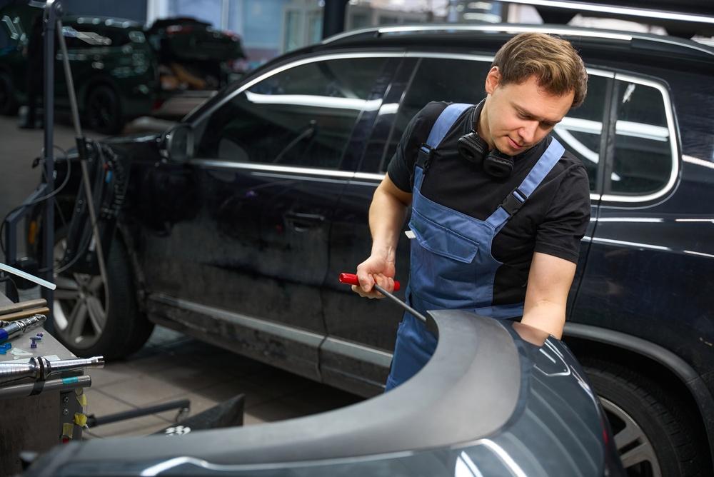 A technician working with an aluminum body panel during car repairs in a body shop bay.